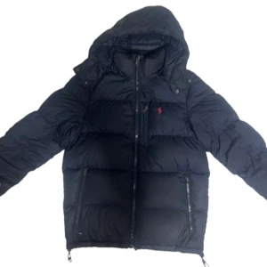 Navy dunjacka från Polo Ralph Lauren - Navy dunjacka från Polo Ralph Lauren med huva och klassisk röd logga på bröstet. Jackan har två dragkedjefickor framtill och quiltad design. Perfekt för kalla dagar med sin fluffiga och varma känsla. Stängs med dragkedja hela vägen upp. Självklart äkta! OBS‼️ ett hål i högra armen (bild 5) kan lätt sy igen men har helt enkelt inte orkat. Därför endast 2000kr men pris kan diskuteras vid snabb affär. 