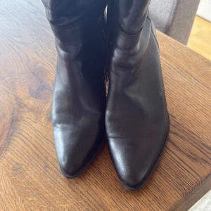 Svarta stövletter med klack - Snygga svarta stövletter i skinn med spetsig tå och kraftig klack på ca 8 cm. Stövletterna har dragkedja på sidan för enkel på- och avtagning. Stilren design som passar till många olika outfits och ger en cool vibe.