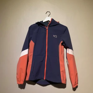 Vindjacka - Kari traa jacka i storlek S. Svårt att få till färgen på bild, men den är lila/rosa. Dragkedja och huva. Material: polyamid och elastan. Finns att hämta i centrala Göteborg eller kan skickas via valfri applikation.