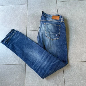 Blå jeans från Tommy Hilfiger, stl 31/32 - Snygga blå jeans från Tommy Hilfiger med klassisk femficksdesign och tydliga slitningar på framsidan. Jeansen har raka ben och normal passform, med Hilfiger-logga på bakfickan och läderpatch i midjan. Perfekt för dig som gillar en avslappnad stil.