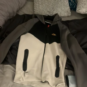 Nike hoodie-jacka i svart, grå och beige - Nike hoodie-jacka med dragkedja i svart, grå och beige. Jackan har huva, två fickor med dragkedja och en liten Nike-logga i guld på bröstet. Perfekt för en sportig och chill stil. Den har inga skador har använts typ 7 gånger 