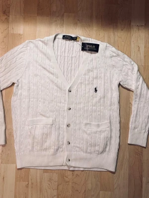 Vit kabelstickad kofta Polo Ralph Lauren - Vit kabelstickad kofta från Polo Ralph Lauren med v-ringning, knappar framtill och två fickor. Klassisk logga broderad på bröstet. Mjuk och skön bomull som passar perfekt till lager på lager. Enkel att styla till olika looks.