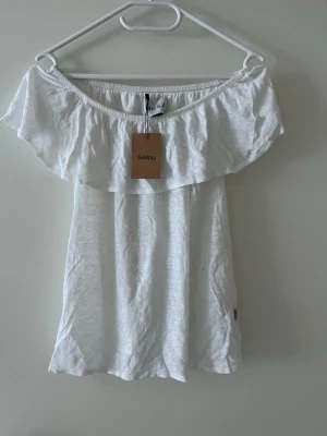 Vit offshoulder topp från elle&il - Säljer en vit offshoulder topp från elle&il i storlek XS. Toppen har en bred volang runt axlarna och är ärmlös med en luftig och lätt passform. Perfekt för varma dagar och enkel att styla med jeans eller kjol.