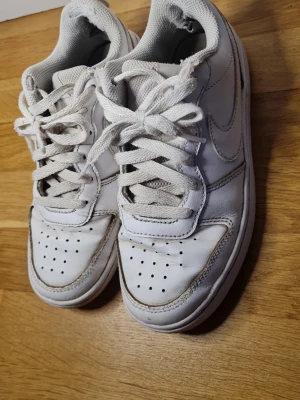 Vita Nike Air Force 1 sneakers - Klassiska vita Nike Air Force 1 sneakers i skinn med perforerad tå och snörning. Swoosh-logga på sidan och vadderad plös i mesh. Perfekta för dig som gillar tidlös streetstyle och vill ha ett par ikoniska sneakers i garderoben.