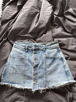Ljusblå jeanskjol/shorts från Folyrose - Säljer en ljusblå jeanskjol/shorts från Folyrose med råa, fransiga kanter och knappar framtill. Kjolen har fickor både fram och bak samt bälteshällor. Perfekt för dig som gillar en avslappnad och trendig denimlook. Det är shorts under kjolen så när du har den på dig och men ser dig bakifrån så ser det ut som shorts. Säljer den då den är för liten❣️