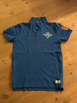 Blå pikétröja Peak Performance - Säljer en blå pikétröja från Peak Performance med tryck på bröstet och siffran 86 i orange på ärmen. Tröjan har klassisk krage med knappslå och orange detalj på insidan av kragen. Perfekt för dig som gillar sportig stil. Nypris 800 kr!
