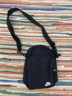 Svart axelväska från Nike - Säljer en stilren svart axelväska från Nike med justerbar rem och två fack med dragkedja. Väskan har en enkel design med Nike-logga framtill och är tillverkad i slitstark polyester. Perfekt för att bära det viktigaste när du är på språng.