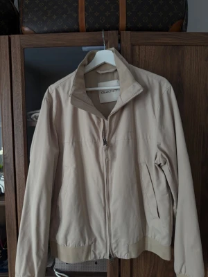Beige vindjacka från GANT - Säljer en stilren beige vindjacka från GANT. Jackan har hög krage, dragkedja framtill och två snedställda fickor. Ribbad mudd vid ärmslut och nederkant ger en clean look. Perfekt för vår och höst när du vill hålla det enkelt men snyggt. Använd två gånger. Storlek M