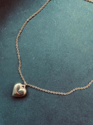 By prisförslag - Guldig halsband med hjärthänge - Snyggt halsband i guldton med ett stilrent hjärthänge. Kedjan är tunn och klassisk, perfekt för att ge en söt touch till din outfit. Hjärtat är slätt och blänkande, vilket gör det enkelt att matcha med andra smycken.