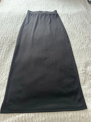 Svart ribbad långkjol med slits - Säljer en svart långkjol med diskret ribbat mönster och slits på sidan. Kjolen har en rak och smal passform som ger en clean look. Perfekt för dig som gillar minimalistisk stil och vill ha något enkelt men ändå snyggt.