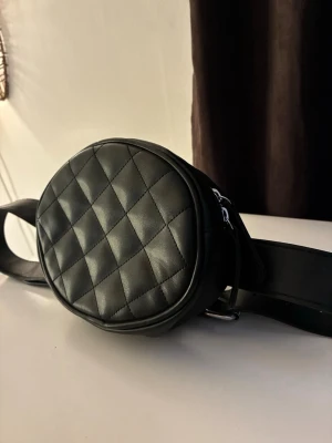 Svart quiltad magväska från Monki - Säljer en svart quiltad magväska från Monki med rund form och bredt justerbart bälte. Väskan har dubbla dragkedjor i silver och är gjord i syntetmaterial som ser ut som skinn. Perfekt för att bära det viktigaste och ger en cool vibe till din outfit.