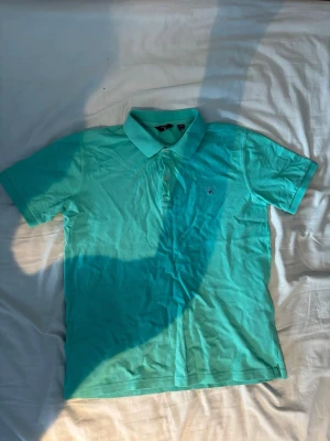 Turkos pikétröja från GANT - Snygg turkos pikétröja från GANT med klassisk krage och knappar framtill. Tröjan har broderad GANT-logga på bröstet och är tillverkad i mjuk bomull. Perfekt till sommaren för en fräsch och sportig look.