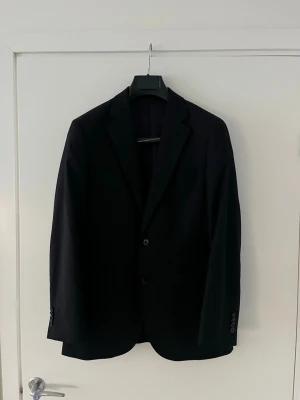 Svart kostym från These Glory Days, strl 48 - Säljer en klassisk svart kostym från These Glory Days i storlek 48. Kavajen har två knappar, smal passform och snygga detaljer vid ärmslutet. Materialet är mjukt och följsamt, perfekt för dig som vill ha en stilren look. Normal passform 