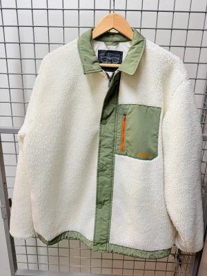 Vit teddyjacka från Levi's med gröna detaljer - Säljer en vit teddyjacka från Levi's med gröna detaljer och orange dragkedja på bröstfickan. Jackan har en grön krage, grönt bröstparti och knappstängning framtill. Materialet är mjuk fleece och syntet, perfekt för dig som gillar chill och streetwear.