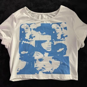Vit croppad t-shirt med blått tryck - Säljer en vit croppad t-shirt i bomull med ett stort blått grafiskt tryck av ansikten på framsidan. T-shirten har korta ärmar och en rund halsringning. Perfekt för dig som gillar unika prints och en avslappnad stil.