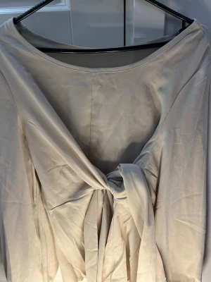 Beige omlottopp med knyt - Säljer en beige omlottopp med lång ärm och snygg knytning framtill. Toppen har en mjuk och slät yta, rundad halsringning och ett elegant fall. Perfekt för dig som gillar stilrena och enkla plagg med en twist.