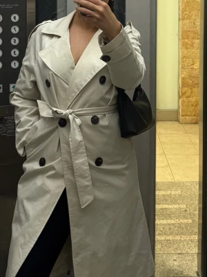 Beige trenchcoat med bälte - Säljer en klassisk beige trenchcoat med dubbelknäppning och bälte i midjan. Jackan har stora slag, svarta knappar och en snygg, lång passform. Perfekt för dig som vill ha en stilren och tidlös look.