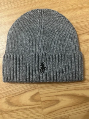 Grå ribbstickad mössa från Polo Ralph Lauren - Säljer en stilren grå ribbstickad mössa från Polo Ralph Lauren med den klassiska svarta loggan broderad framtill. Mössan har vikt kant och är perfekt för att hålla värmen under kyliga dagar. Enkel att matcha med olika outfits.