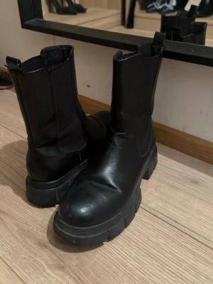Chunky svarta chelsea boots - Säljer ett par chunky svarta chelsea boots i skinn med elastiska paneler på sidorna och grov platt sula. Bootsens höga skaft och minimalistiska design gör dem perfekta till jeans eller kjol. Lätta att dra på tack vare öglan bak.