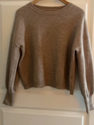 Beige stickad tröja med ribbade muddar - Mysig beige stickad tröja med rund hals och ribbade muddar vid ärmslut och nederkant. Tröjan har en relaxed passform och är perfekt för lager på lager under kyliga dagar. Enkel och stilren design som passar till det mesta.