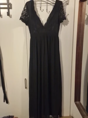 Svart långklänning med spetsdetaljer - Säljer en svart långklänning med v-ringning och korta spetsärmar. Överdelen är dekorerad med vacker spets och kjolen faller mjukt. Klänningen har dragkedja i ryggen och är perfekt för dig som vill ha en elegant look.