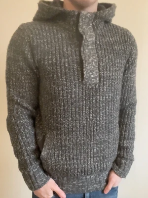Grå stickad hoodie - Otroligt snygg, varm och skön grå stickad hoodie med knappar vid halsen. Passar perfekt nu till vintern. Priset kan såklart diskuteras! Modellen är 180cm, 75kg.