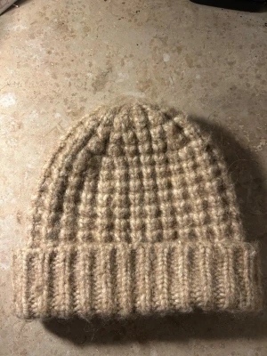 Beige stickad mössa från & Other Stories - Säljer en tjock stickad mössa i beige från & Other Stories. Mössan har en bred uppvikt kant och är gjord i ett mjukt, fluffigt garn som ger en mysig känsla. Perfekt för kalla dagar och enkel att matcha med olika outfits.