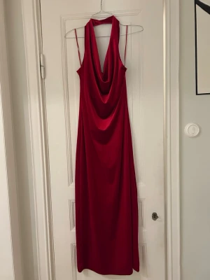 Röd långklänning med draperad hals - Säljer en röd långklänning i mjukt, glansigt material med draperad halsringning och tunna axelband. Klänningen har en snygg halterneck och figurnära passform som ger en elegant siluett. Perfekt för dig som vill sticka ut med en klassisk look. Använd endast en gång