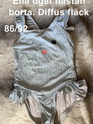 Randig baddräkt med volanger - Söt baddräkt med ljusblå och vita ränder, volangdetaljer vid axlar och höfter samt ett gulligt ansikte framtill. Perfekt för strand eller poolhäng. 🌸 Ena ögat är nästan borta. Diffusa fläckar. Storlek 86/92