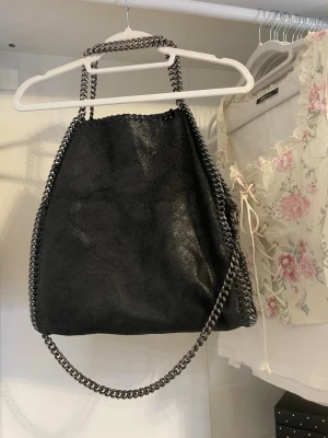 Svart axelväska med kedjedetaljer - Axelväska med silverfärgade kedjor som handtag och kantdetalj. 