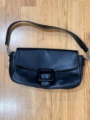 Svart axelväska med stor spänne - Snygg svart axelväska i slätt skinnliknande material med en bred axelrem och ett stort dekorativt spänne på framsidan. Väskan har en stilren och minimalistisk design som passar till många olika looks.