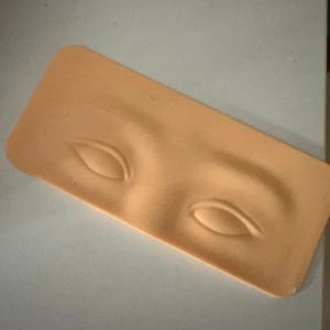 Övningsplatta för ögonsmink - Säljer en beige övningsplatta i mjukt silikonmaterial med formade ögon och bryn. Perfekt för att träna på att lägga ögonskugga, eyeliner eller ögonbryn. Plattan är rektangulär och har en realistisk hudtonad färg.