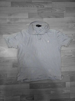 Randig piké från Polo Ralph Lauren - Ljusgrå pikétröja från Polo Ralph Lauren med tunna vita ränder och klassisk krage. Ikoniska broderade loggan på bröstet. Tillverkad i mjuk bomull och har korta ärmar. Perfekt för en avslappnad och stilren look.