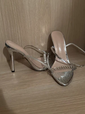 Silvriga klackar med strassdetaljer - Säljer ett par riktigt snygga silvriga klackar med krokodilpräglad yta och transparenta remmar dekorerade med strass. Skorna har spetsig tå och hög klack, perfekta för att sticka ut. Materialet är syntet och de har en glittrig finish.