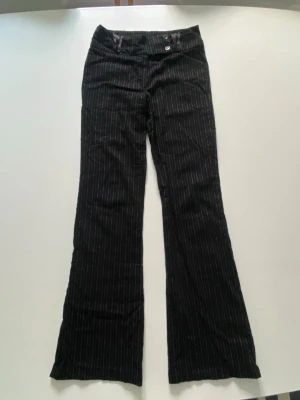 Svarta randiga bootcut byxor - Säljer ett par svarta kostymbyxor med tunna vita ränder och bootcut-modell. Byxorna har låg midja och klassisk stängning med knapp och dragkedja.