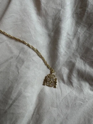 Guldig halskedja med Spaniens symbol  - Säljer en snygg guldig halskedja med ett detaljerat Spanien symbol. Hänget har glittrande stenar och en krona upptill, samt två pelare på sidorna. Kedjan är tvinnad och har justerbar längd med klassisk låsning. Perfekt accessoar för att lyfta din outfit.