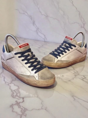 Crime London sneakers  - Säljer ett par vita sneakers från Crime med blå snörning och blå häl. Skorna har texten 'DISTRESSED WITH NO STRESS' på sidan och en beige sula. Materialet är skinn och de har rund tå och platt sula. Perfekta för dig som gillar stilrena och coola sneakers. hör av dig vid minsta fundering och jag fraktar alltid en till två dagar efter beställningen! Pris kan diskuteras vid snabb affär😊