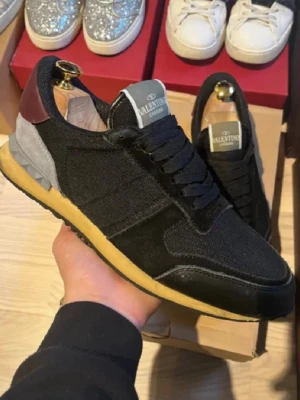 Valentino Garavani svarta sneakers - Svarta Valentino Garavani sneakers med detaljer i grå mocka och vinröd häl. Det finns lite små defekter men inget som är värsta och skadar skorna Ovandel i textil och skinn, klassisk snörning och gulaktig sula. Snygg och sportig design med diskret logga på plösen. Perfekta för dig som vill ha en exklusiv touch på din streetstyle.
