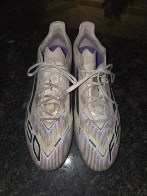 Adidas F50 Elite FG Road to Glory - Säljer ett par Adidas F50 Elite FG Road to Glory fotbollsskor i vitt, svart och guld. Skorna används av Lionel Messi, Lamine Yamal, Florian Wirtz och Trinity Rodman Den lätta Fibertouch-ovandelen på dessa skor är skapad i distinkta färger och en Sprintweb 3D-struktur säkerställer att varje touch är minnesvärd Den konstruerade textilen ger en anpassningsbar men stödjande passform, och det taktila övre höljet ger optimal känsla av bollen vid höga hastigheter En avancerad yttersula kallad Sprint