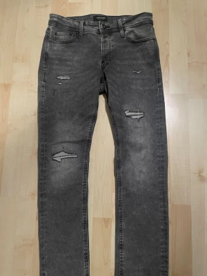 Grå slitna slim jeans Jack & Jones - Säljer ett par grå slim fit jeans från Jack & Jones, modell Glenn. Jeansen har slitna detaljer och hål på benen för en riktigt edgy look. Klassisk femficksmodell med knappgylf och smal passform. Perfekta till sneakers och hoodie. Storlek W30 L30