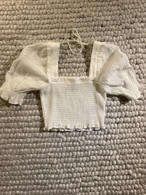 Puffärmad blus från H&M - Supersöt vit blus från H&M i storlek XS. Den har puffiga korta ärmar och smockad kropp. Fyrkantig urringning, volangkant nedtill och tunna knytband i nacken. Tunn och luftig, perfekt för varma dagar. Några små bruna prickar i kragen på baksidan, men har aldrig sett de förens nu när jag tog bilderna. Såljer pga att den inte kommer till användning för mig.