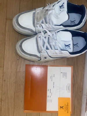Louis Vuitton LV Trainer sneakers vit/blå - Säljer ett par Louis Vuitton LV Trainer sneakers i vitt och blått skinn med grå detaljer. Klassisk LV-logga på plösen och sidan, samt #54 broderat bak. Skorna har snörning, platt sula och rund tå. Kommer med originalbox och certifikat. Använd 3 gånger kvitto finns storlek 45