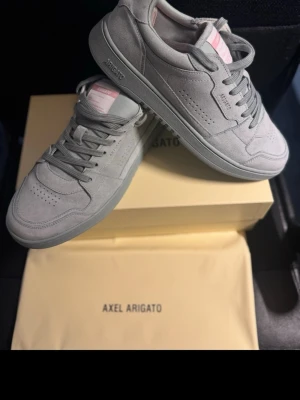 Grå sneakers från Axel Arigato 43 mocka - Snygga grå sneakers från Axel Arigato i mjuk mocka med perforerade detaljer på tån och stilrena linjer. Skorna har matchande grå snörning, rosa innerfoder och subtil branding på sidan och hälen. Perfekta för dig som gillar minimalistisk och modern stil.