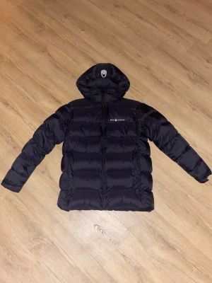 Svart pufferjacka från Sail Racing/S - Säljer en svart pufferjacka från Sail Racing med huva och dragkedja framtill. Jackan har en quiltad design och är tillverkad i syntetmaterial. Perfekt för kalla dagar och har en clean look med diskret logga på bröstet.Strorlek S.