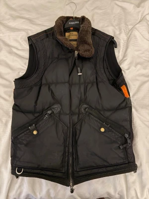 Svart dunväst från Parajumpers - Säljer en svart dunväst från Parajumpers med fluffig brun krage, två stora snedställda fickor med dragkedja och knapp, samt orange meshdetaljer på insidan. Västen har dubbel dragkedja framtill och coola detaljer som metallring och patch bak i nacken.