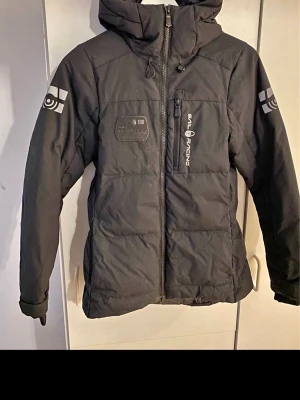 Svart pufferjacka Sail Racing - Säljer en svart pufferjacka från Sail Racing med huva och coola loggor på bröstet och ärmarna. Jackan har dragkedja framtill, flera fickor och justerbara muddar. Perfekt för kalla dagar och riktigt snygg streetstil.