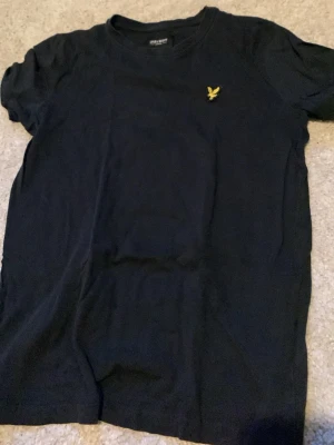 Svart t-shirt från Lyle & Scott Junior - Klassisk svart t-shirt från Lyle & Scott Junior med gul broderad logga på bröstet. Enkel och stilren design med rund hals och korta ärmar. Tillverkad i mjuk bomull för skön känsla hela dagen. Pris kan diskuteras 