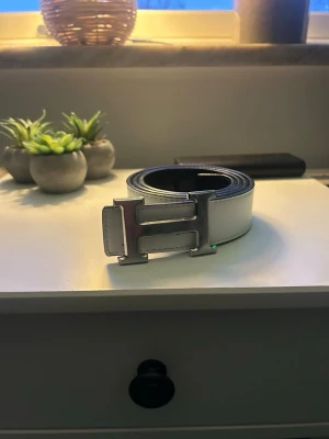 Vändbart vitt och svart bälte med H-spänne - Säljer ett stilrent vändbart bälte i vitt och svart med ett stort H-format metallspänne. Bältet är i slätt material på den vita sidan och har en mer texturerad yta på den svarta sidan. Perfekt accessoar för att lyfta din outfit.
