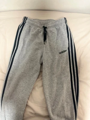 Grå mjukisbyxor från Adidas M - Säljer ett par grå mjukisbyxor från Adidas i storlek M. Byxorna har svarta klassiska ränder längs sidorna, snörning i midjan och Adidas-logga på benet. Tillverkade i mjukt bomullsmaterial, perfekta för chill eller träning.
