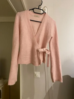 Rosa omlottkofta från Gina Tricot - Supermjuk och fluffig rosa omlottkofta från Gina Tricot i storlek XS. Koftan har långa ärmar, v-ringning och knytband i sidan. Perfekt för lager på lager och ger en söt och mysig vibe till din outfit.